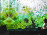 A woman sits serenely in a room with large format digital display casting a digital seascape scene